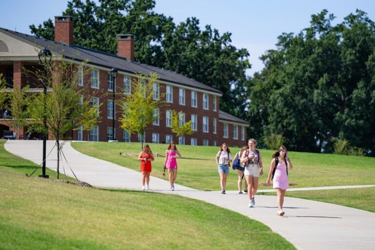 Students walk down a sidewalk and away from a residence hall on a sunny day on a college campus.