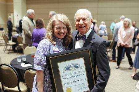A white woman with light hair and a white older man with a bowtie. The man holds a commendation. Harry Shucker