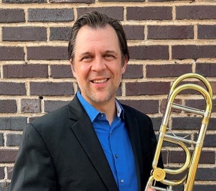 A white man in a sport coat holds a trombone and stands against a brick wall. Cory Mixdorf
