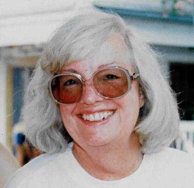 A white woman with white hair wears sunglasses in an outdoor photo. Judy Bainbridge.