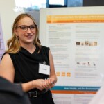 A blonde white woman wearing glasses stands in front of a research poster talking to people we can't see.