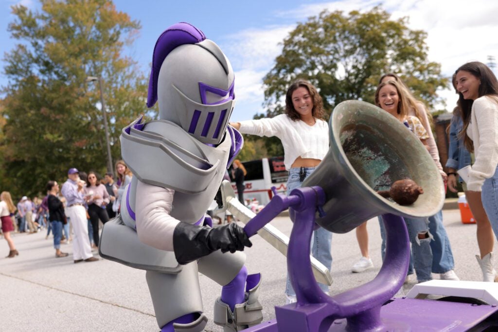 paladin mascot ringing large bell, outdoors, blue sky with clouds, green trees