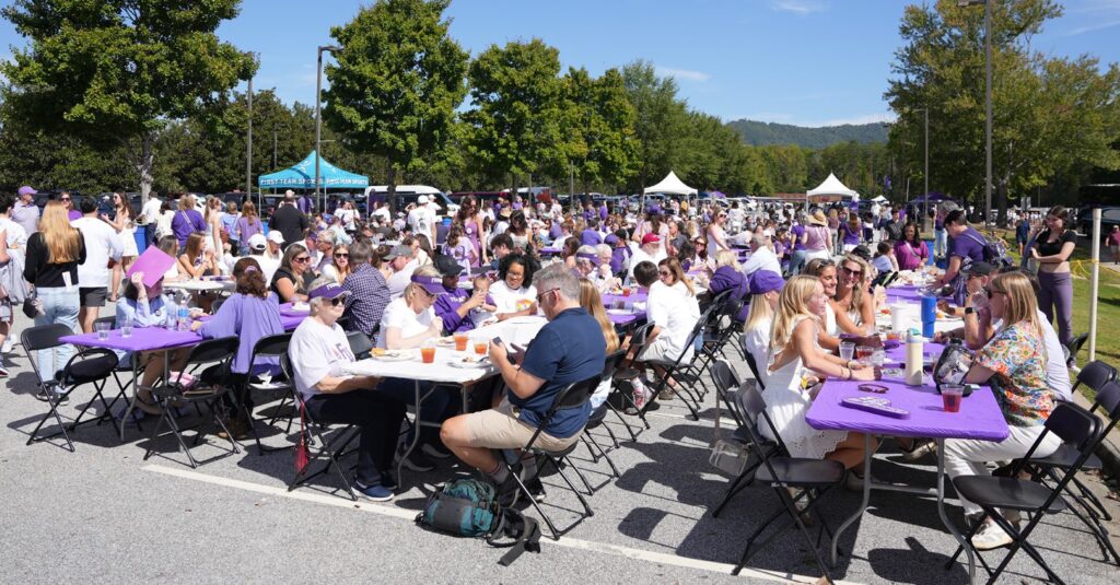 picture of a large crowd seated at tables for a BBQ out in the sun. Tables are covered with purple cloths.