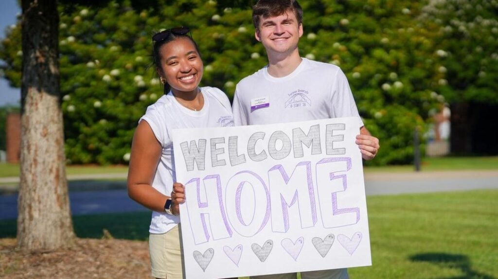 a young woman and young man stand together holding a sign that reads "Welcome Home" with a line of purple hearts below the text. This was part of Summer Orientation 2026.