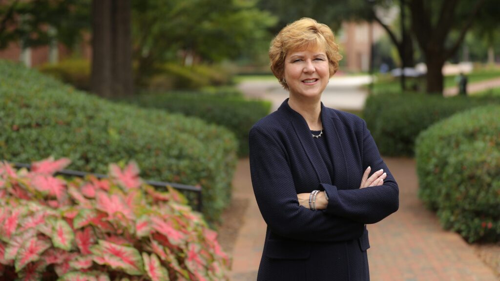 President Elizabeth Davis stands on a brick walkway with hedges behind her and a red leafed plant to her right. She is wearing a black blazer and her arms are crossed. She faces the camera and smiles.