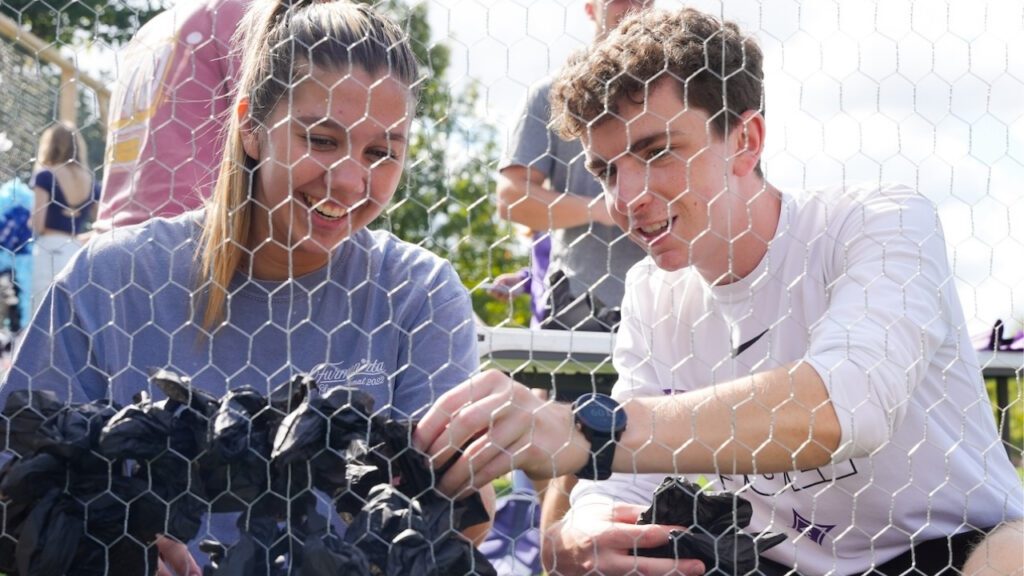Two students sit together (woman on the left, man on the right). There is chicken wire between them and the camera lens and they are in the process of "pomping" which is to add tissue paper into the holes of the chicken wire, from which parade or Homecoming floats are built.