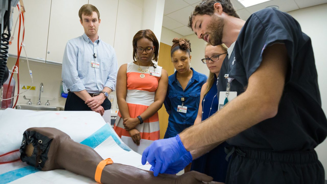 Students watching a nurse take blood from a dummy arm
