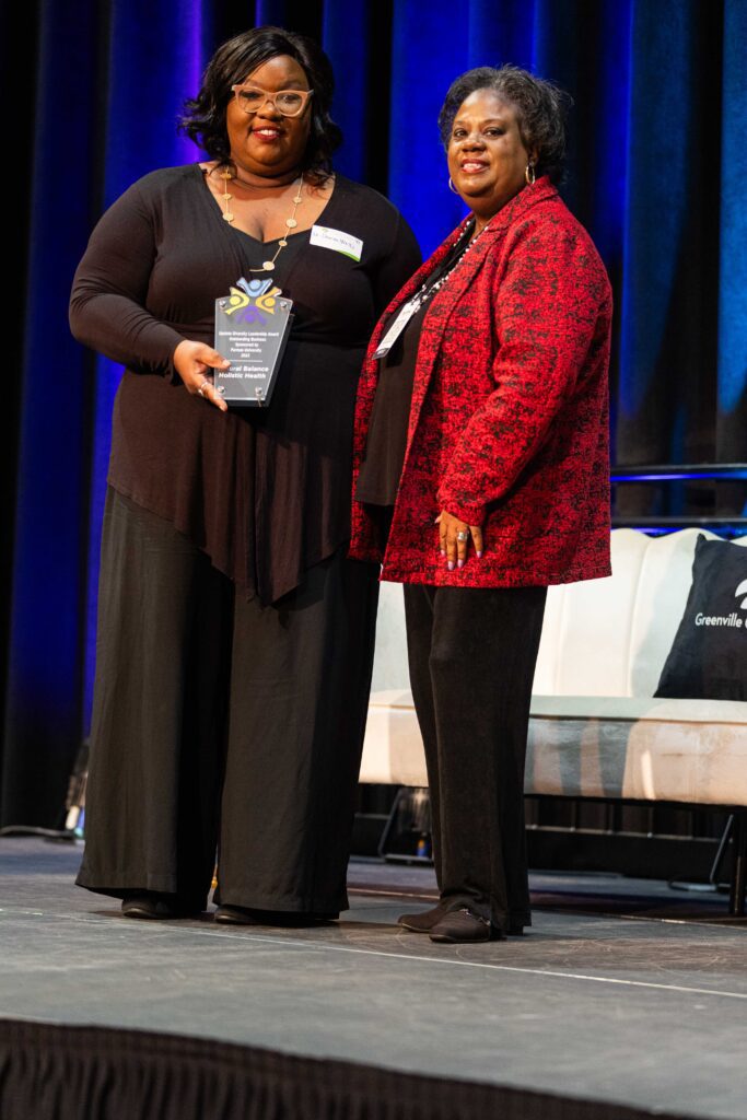 Two women standing on stage. Woman on the left holds an award.