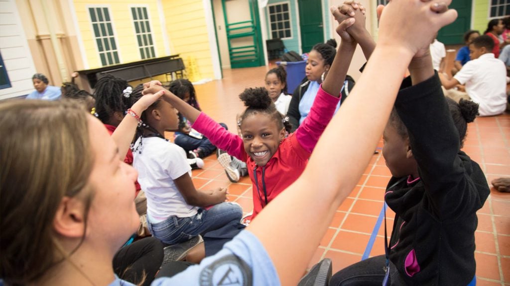 A group of children sit in a circle, grasping each other's hands and holding them up in the air in an activity led by a counselor