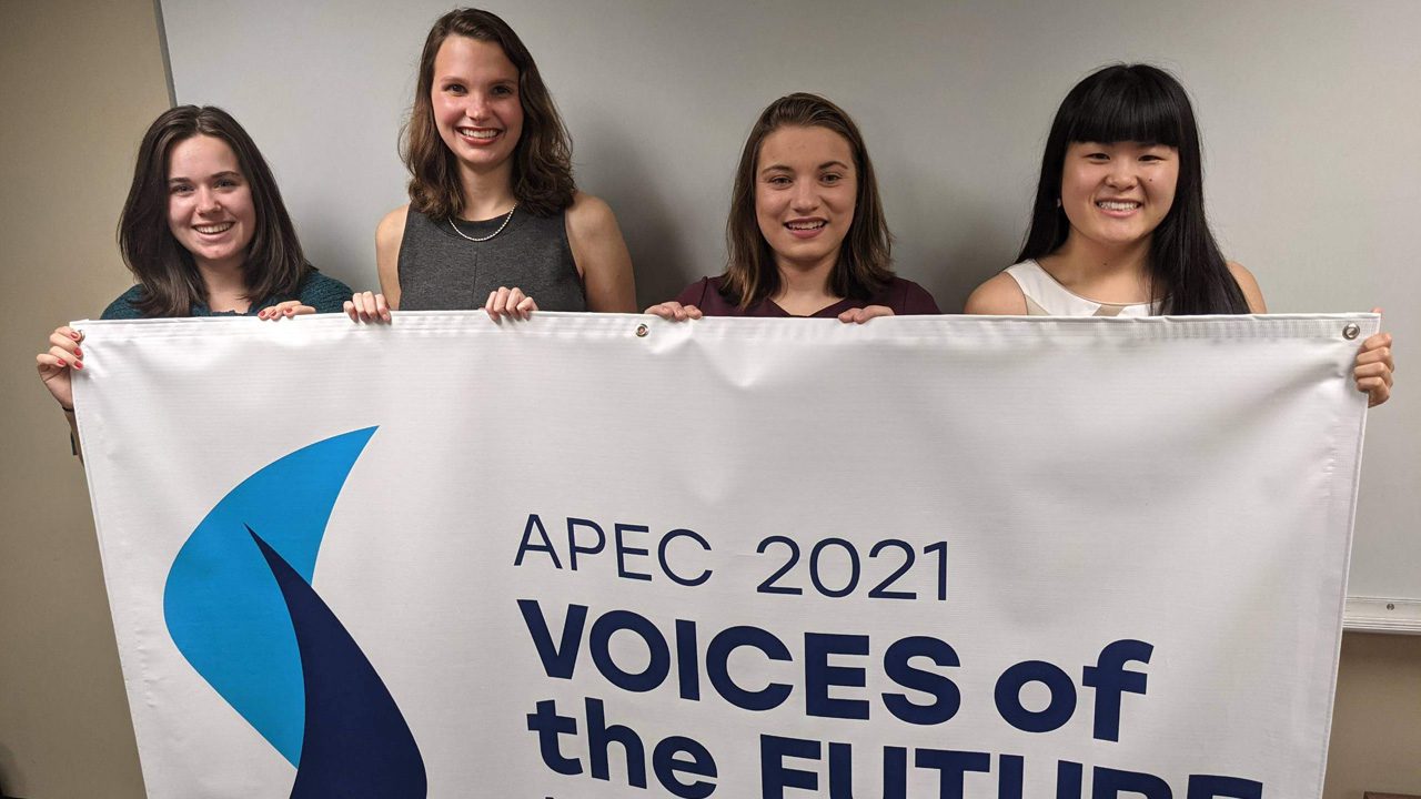 Delegates Erica Daly, Lauren Garrison, Grace Curty, and Megan Litz pose in front of an APEC banner ahead of the first virtual session