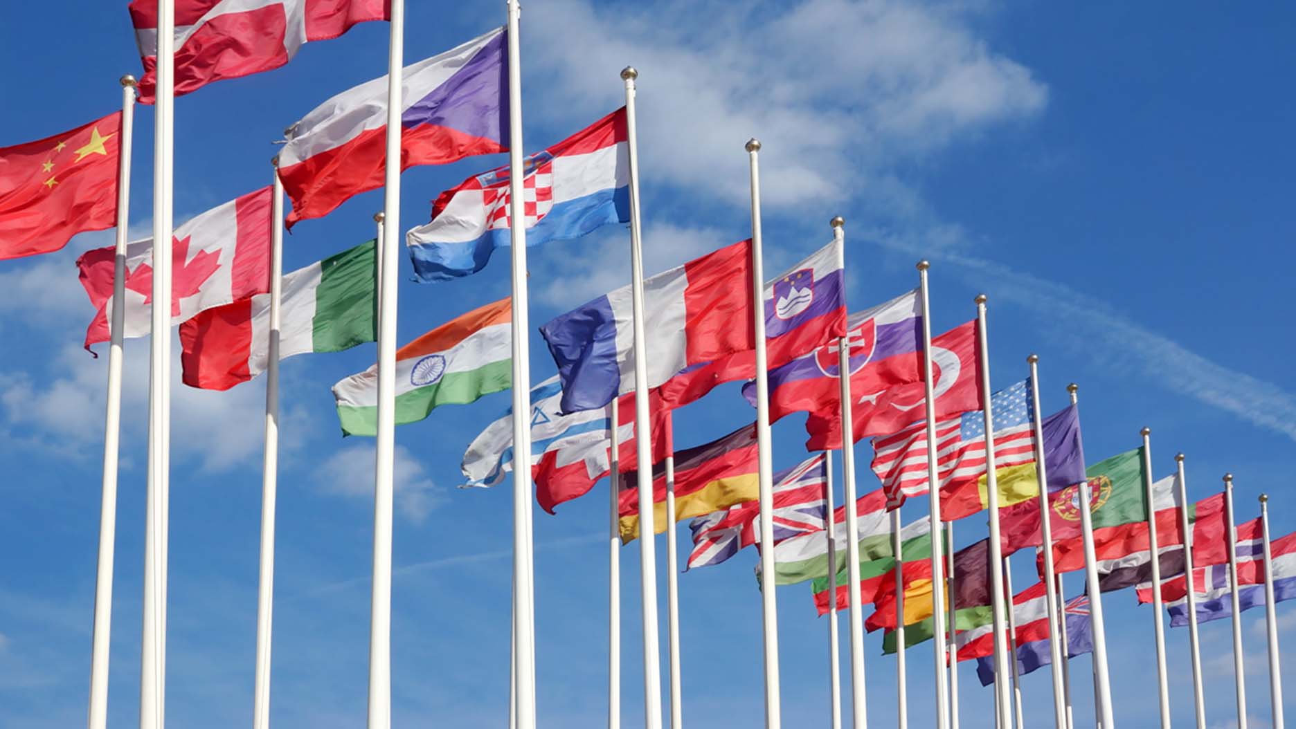 An array of flags from various countries, each positioned on its own flag pole. They stand in front of a bright blue sky.