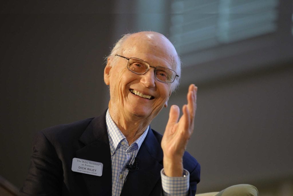 Secretary Dick Riley sits on stage at an event. He is wearing a black suit, checkered shirt, and glasses. He is smiling and gesturing with his hand toward fellow panelists.