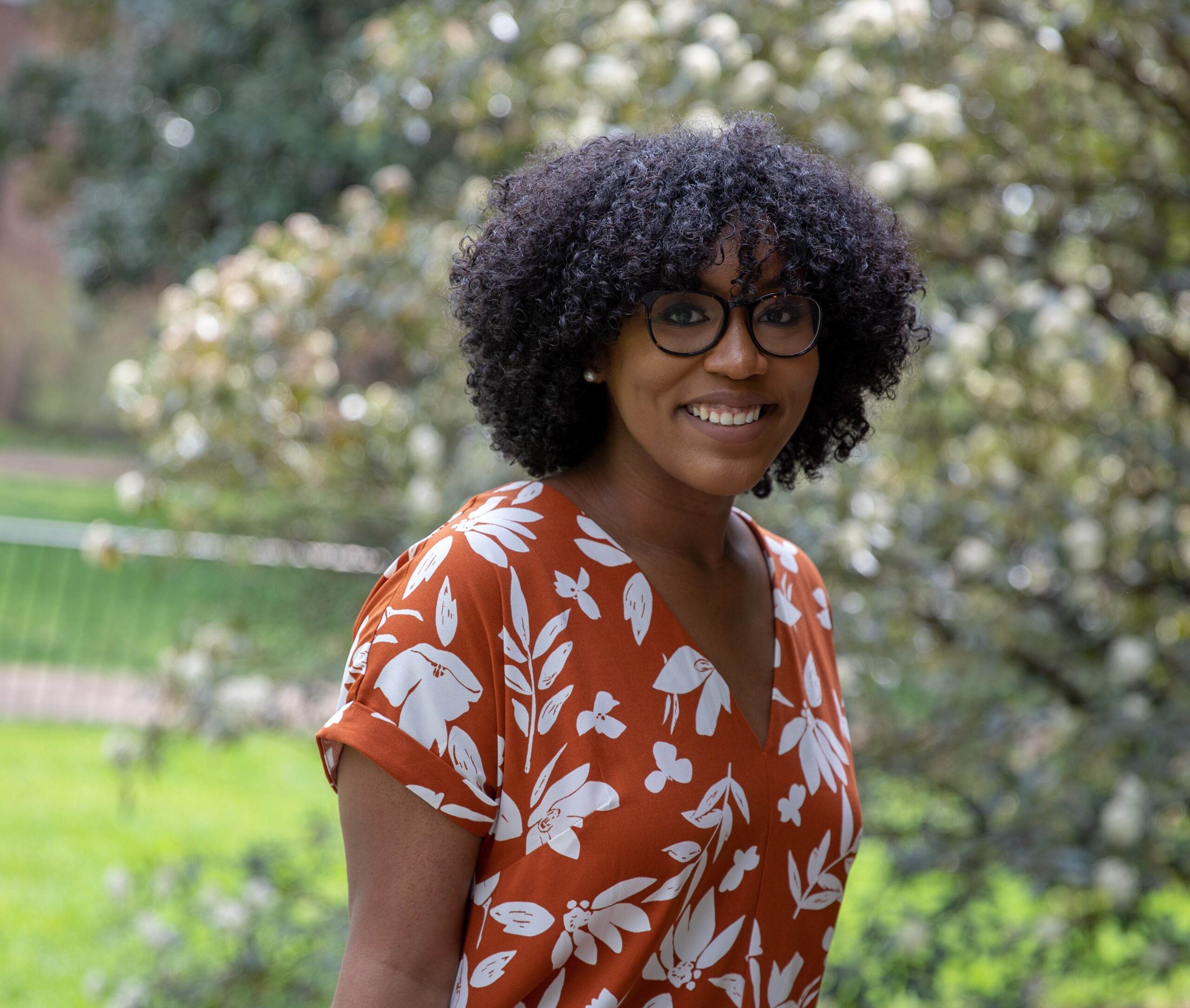 Jocelyn is a Black woman with curly hair who is standing outdoors at a university. She is smiling and wearing an orange dress.
